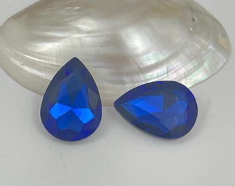 以下が含まれることがあります： 深いコバルト色の洋ナシ型でファセットカットされた青いガラスのカボションが2つ。石は尖った上部と丸みを帯びた基部を持ち、光を反射する幾何学的なファセットが特徴です。これらの鮮やかな青い宝石は、貝殻の前に配置されています。