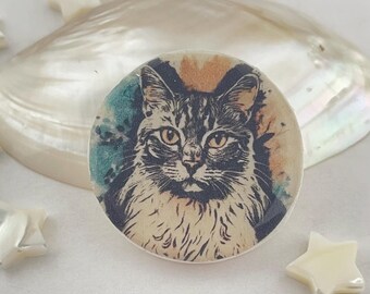 以下が含まれることがあります： 水彩画の背景を持つ丸い猫の肖像画。猫はグレーとタンの色合いで描かれ、詳細な毛皮と琥珀色の目があります。背景には、ティールとオレンジの色合いがあります。装飾的な貝殻と星型のアクセントが肖像画を囲んでいます。