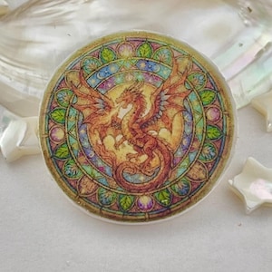 May include: Round, stained-glass style dragon design with a gold border. The dragon is depicted in shades of orange and brown, with outstretched wings. The background features a mix of green, blue, and yellow hues.