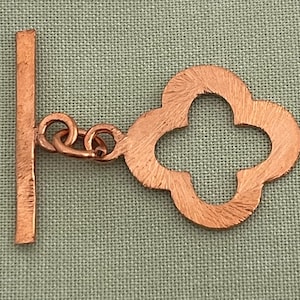 May include: A copper-colored toggle clasp with a clover-shaped end and a rectangular bar. The two pieces are connected by small, round links. The clasp has a textured surface and is set against a muted green background.