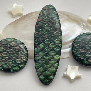 May include: A set of three iridescent green and black resin jewelry pieces with a scale pattern. The pieces include a long oval pendant, a round pendant, and a smaller round pendant.