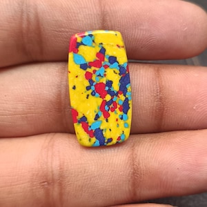 Könnte beinhalten: Ein rechteckiger Cabochon-Edelstein mit einer leuchtend gelben Grundfarbe und einem gesprenkelten Muster aus leuchtend roten, himmelblauen und dunkelblauen Punkten, die über die glatte, polierte Oberfläche verteilt sind.