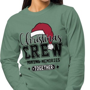 May include: Sage green long-sleeve top featuring the text "Christmas CREW MAKING MEMORIES TOGETHER" in black and white, with a Santa hat graphic. The top is made of a soft material and is suitable for the festive season.