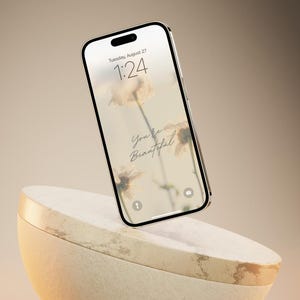 May include: A white smartphone with a floral wallpaper and the text "You're Beautiful" displayed on the screen. The phone is floating above a beige, textured, half-sphere.
