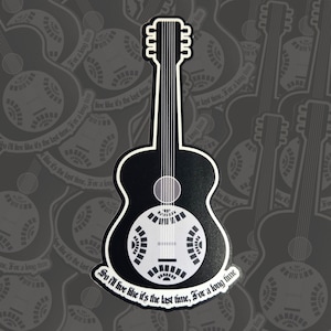 May include: A black and white sticker featuring a resonator guitar design. The guitar has a distinctive round sound hole and a stylized appearance. The text "So I'll live like it's the last time, For a long time" is written on the bottom.