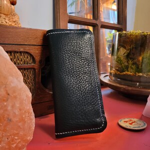Peut inclure: Un étui de téléphone en cuir noir avec des coutures blanches. L'étui a une surface texturée et est posé sur une surface rouge.