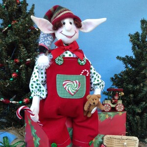 May include: A Christmas elf doll wearing a red and green outfit with a candy cane in its hand. The elf is sitting on a red and green gift box with a teddy bear in its hand. The elf is surrounded by Christmas decorations, including a Christmas tree and a toy train.