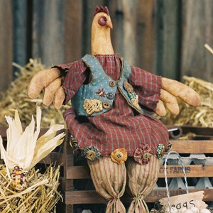 Può includere: Una statuina di gallina fatta a mano con corpo in tessuto, che indossa un gilet e un vestito a quadri. La gallina ha lunghe zampe di legno ed è appollaiata su una cassa di legno. Un cestino di filo di ferro con uova e mais è nelle vicinanze, creando un tema rustico da fattoria.