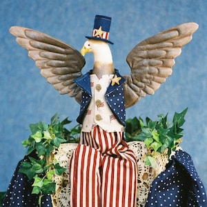 Könnte beinhalten: Eine patriotische Adlerpuppe, die einen blauen Zylinder mit einem Stern, eine blaue Weste mit weißen Sternen und eine rot-weiß gestreifte Hose trägt. Der Adler sitzt auf einer weißen Häkelspitze mit blauem Stoff mit weißen Sternen dahinter.