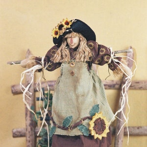 May include: A handmade scarecrow doll with a brown hat, a green and white checkered dress, and a sunflower on the side. The doll is standing on a bed of straw with fall foliage.