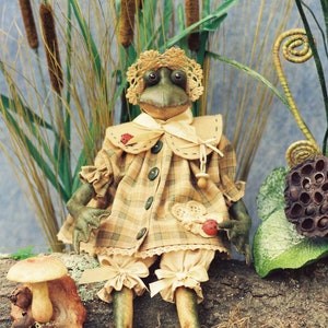 May include: A whimsical frog doll with a plaid dress, lace-trimmed bloomers, and a bonnet. The doll is seated on a log with a mushroom and cattails in the background. The frog has green skin and large, expressive eyes.