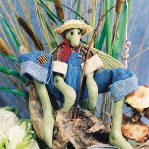 May include: A green grasshopper doll wearing a straw hat, blue overalls, and a red and white checkered shirt is sitting on a piece of driftwood holding a fishing pole with a worm on the line. The doll is surrounded by greenery and a small metal bucket.