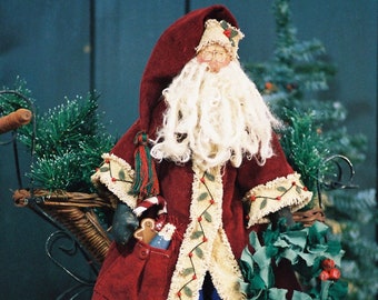 May include: A handmade Santa Claus doll wearing a red coat with white trim, blue pants, and a white beard. He is holding a wreath and a bag of toys. The doll is sitting on a crocheted doily.