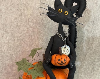 May include: A black cat figurine with yellow eyes and a silver chain necklace, holding a small orange pumpkin. The cat is seated on a larger orange pumpkin with green felt leaves. A tag on the necklace reads "Boo". The figurine is on a wooden base.