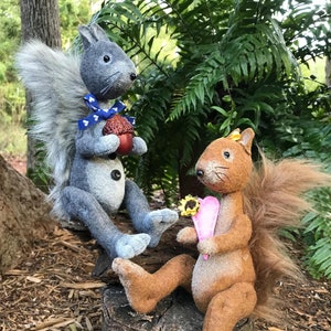 May include: Two plush squirrel figurines sitting on a tree stump. The gray squirrel is holding an acorn and wearing a blue bow tie. The brown squirrel is holding a pink heart and a sunflower.