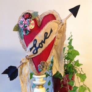 May include: A red felt heart with a lace trim and a banner that says "Love" is pierced by a black arrow. The heart is on a white pedestal with a gold base. A green plant is in a red pot behind the heart.