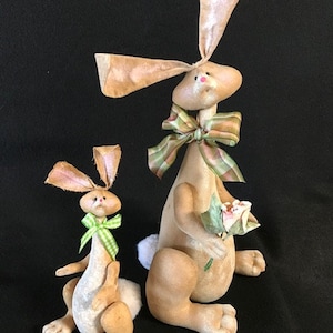 Könnte beinhalten: Zwei handgefertigte Stoffhasenfiguren. Der größere Hase hält einen Blumenstrauß und trägt eine grün-braune Karobinde. Der kleinere Hase trägt eine grün-weiß gestreifte Schleife. Beide Hasen sind braun und weiß.