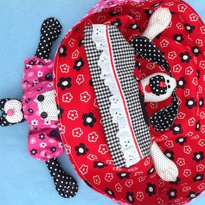 May include: A black and white polka dot dog toy with red and white floral fabric lining. The toy is lying in a red and white floral fabric bed.