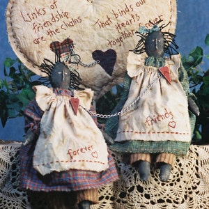 Könnte beinhalten: Zwei Stoffpuppen mit schwarzen Gesichtern und schwarzen Haaren sind durch eine Kette mit Herzen verbunden. Die Puppen tragen karierte Kleider und Schürzen. Die Puppen sitzen auf einer weißen Häkelspitze. Der Hintergrund ist eine blaue Wand mit einem herzförmigen Stoffbanner, auf dem steht "Links of friendship are the chains That binds our hearts together forever".
