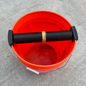 May include: An orange plastic bucket with a black PVC pipe handle attached to the rim. The pipe is secured with black plastic end caps and brown tape.