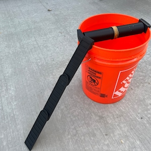 May include: An orange plastic bucket with a black and gold pipe and a black plastic ladder-like tool. The bucket has a metal handle and a warning label. The tool is resting on the bucket's rim.