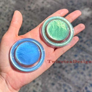 Może przedstawiać: Dwa iryzujące, okrągłe, dekoracyjne przedmioty w odcieniach niebieskiego i zielonego. Obiekty mają przezroczystą, okrągłą obw&oacute;dkę i błyszczący, teksturowany środek. Widoczny jest tekst "TwiceteaDesign".