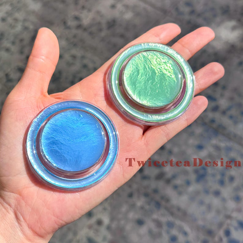 Może przedstawiać: Dwa iryzujące, okrągłe, dekoracyjne przedmioty w odcieniach niebieskiego i zielonego. Obiekty mają przezroczystą, okrągłą obw&oacute;dkę i błyszczący, teksturowany środek. Widoczny jest tekst "TwiceteaDesign".