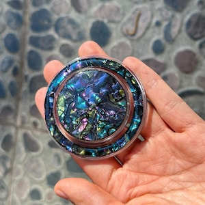 May include: A circular abalone shell coaster with a clear plastic edge. The coaster features a shimmering display of blues, greens, and purples. The abalone shell has a natural, textured look. The coaster is held in a hand.