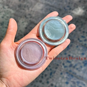 Może przedstawiać: Dwa okrągłe, przezroczyste pojemniki z iryzującą zawartością trzymane w dłoni. Jeden zawiera blador&oacute;żowy, drugi blękitny. Widoczny jest tekst "TwiceteaDesign". Pojemniki mają podniesioną krawędź i wydają się być produktami kosmetycznymi.