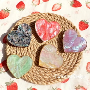 May include: Five heart-shaped decorative pieces with iridescent surfaces and gold-coloured trim. The hearts are in shades of green, pink, red, and blue, and are arranged on a woven rope mat. The background features a strawberry pattern.