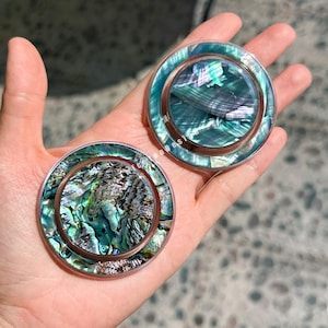 May include: Two round, iridescent abalone shell phone grips with a clear border and copper-coloured ring. The abalone shells display shades of green, blue, and silver. The grips are held in a person's hand.