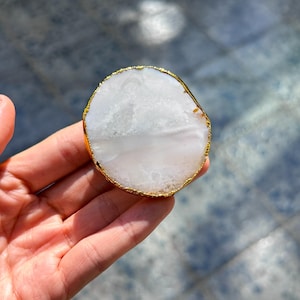 Può includere: Una fetta di agata bianca con un bordo dorato. La pietra naturale ha una superficie lucida ed è tenuta in mano. L'agata ha un motivo unico ed è un oggetto decorativo.