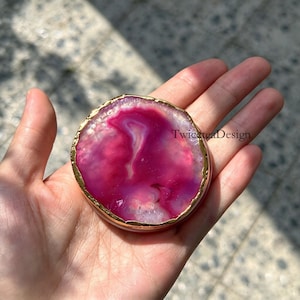 Könnte beinhalten: Eine kreisförmige Scheibe aus poliertem Achatstein mit leuchtenden Magenta- und Rosatönen sowie durchscheinenden kristallinen Mustern. Die Kanten des Steins sind mit einem metallisch goldenen Rand versehen. Der Text TwiceTeaDesign ist sichtbar.