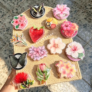 May include: A collection of circular phone grips featuring various designs including a black and yellow butterfly, a pink lily, a red strawberry, a slice of citrus fruit, several floral patterns in shades of pink, red, and yellow, and a grey butterfly.