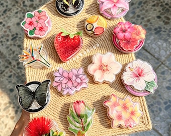May include: A collection of circular phone grips featuring various designs including a black and yellow butterfly, a pink lily, a red strawberry, a slice of citrus fruit, several floral patterns in shades of pink, red, and yellow, and a grey butterfly.