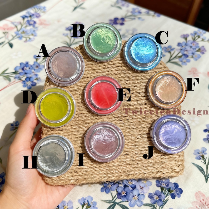 Może przedstawiać: Kolekcja dziesięciu okrągłych, iryzujących pojemnik&oacute;w kosmetycznych w r&oacute;żnych kolorach, w tym r&oacute;żowym, zielonym, niebieskim, ż&oacute;łtym i srebrnym. Pojemniki są ułożone na tkanej powierzchni z juty, z kwiatowym tłem.