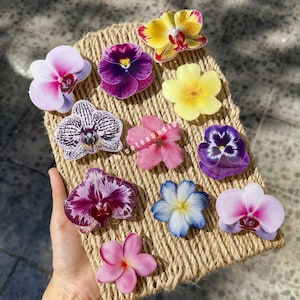 Op de afbeelding: Een verzameling kleurrijke bloemvormige harsstukken, waaronder orchideeën, viooltjes en hibiscus, gerangschikt op een geweven jute mat. De bloemen tonen een verscheidenheid aan kleuren, zoals roze, paars, geel en wit, met ingewikkelde details.