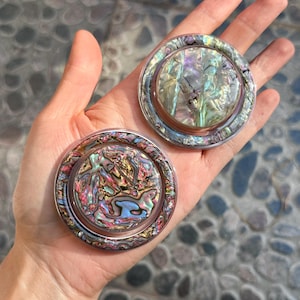 May include: Two round abalone shell coasters with a clear, raised edge. The coasters display a swirling pattern of pink, blue, green, and gold. The coasters are held in a hand. The coasters are approximately 10 cm in diameter.