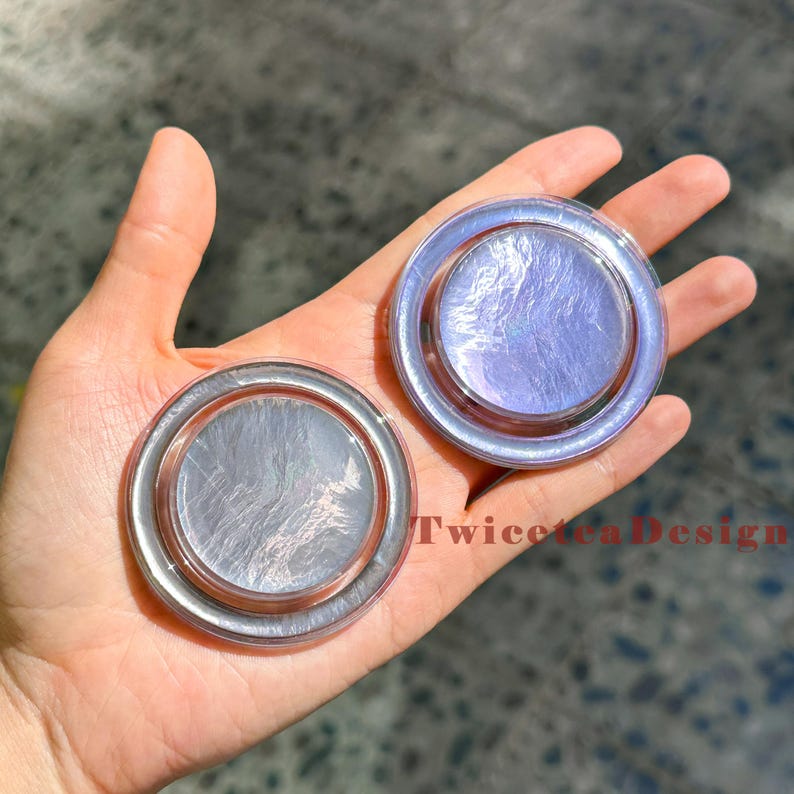 Może przedstawiać: Dwa iryzujące, okrągłe, dekoracyjne przedmioty w srebrno-fioletowej kolorystyce. Obiekty mają okrągły kształt z podniesioną krawędzią i odblaskową, teksturowaną powierzchnią. Widoczny jest tekst "TwiceTeaDesign".