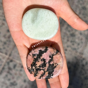 Könnte beinhalten: Zwei polierte Steine: einer hellgrün, der andere rosa mit schwarzen Adern. Die Steine sind oval und glatt. Der Text "Twicetea" ist auf dem Bild sichtbar. Diese Steine werden wahrscheinlich für Meditation oder Heilung verwendet.