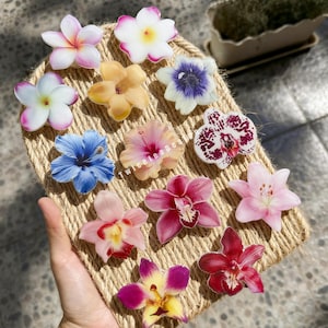May include: A collection of colorful flower-shaped accessories, including plumeria, hibiscus, and orchids. The flowers are in shades of pink, yellow, blue, and white. The accessories are arranged on a woven, rope-like surface.