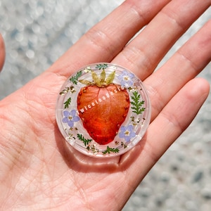 Puede incluir: Un objeto redondo y transparente de resina con una fresa roja vibrante en el centro. La fresa está rodeada de pequeñas flores azules, hojas verdes en forma de helecho y motas doradas. El texto "twiceteadesign" es visible.