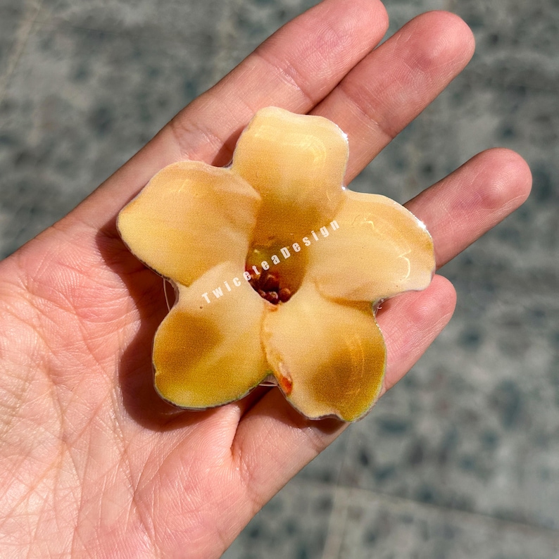 May include: A yellow flower-shaped phone grip with a brown center. The phone grip has the text "Twiceteadesign" on it. The flower has five petals and is held in a hand.