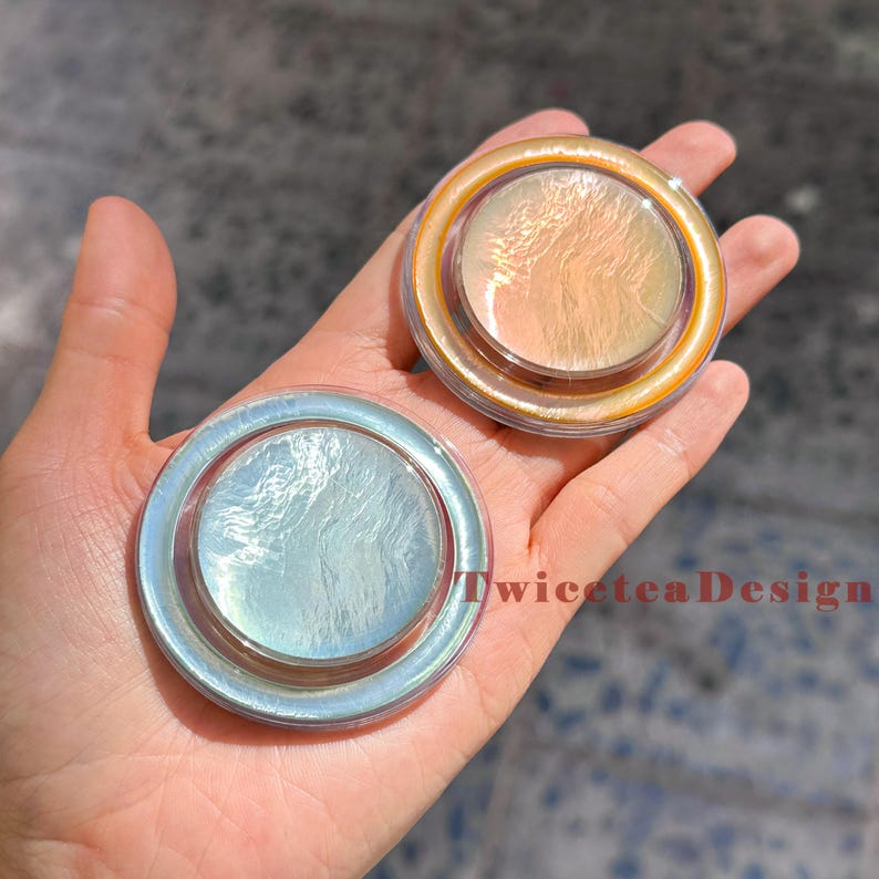 Może przedstawiać: Dwa iryzujące, okrągłe kompakty z perłowym połyskiem trzymane w dłoni. Jeden jest w kolorze jasnoniebieskim, a drugi w ciepłym pomarańczowym. Tekst "TwiceteaDesign" jest widoczny na zdjęciu.