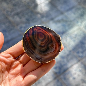 Può includere: Una fetta di pietra d'agata lucidata con un bordo color oro. La pietra presenta bande concentriche di tonalità marroni, nere e rosso-arancio. La pietra naturale è tenuta in una mano, mostrando i suoi motivi e le sue trame uniche.