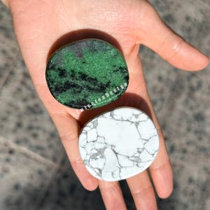 Könnte beinhalten: Zwei polierte Steine, einer grün mit schwarzen Einschlüssen und der andere weiß mit grauer Maserung, werden in einer Hand gehalten. Der grüne Stein trägt den Text "twiceteadesign". Die Steine sind oval und glatt.