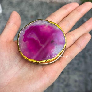 May include: A vibrant, round coaster with a gold-colored edge. The coaster features a striking magenta and white agate pattern. The design is natural and organic, with a polished finish. The text "Twicet design" is visible on the side.