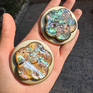 May include: Two gold-coloured, round decorative objects with a four-leaf clover design. Each object's centre has iridescent abalone shell in blue, green, and brown. The objects are held in a hand.