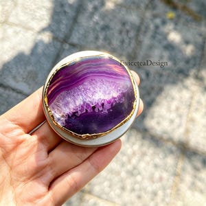 Könnte beinhalten: Ein runder Handyhalter mit einer polierten violetten Achat-Scheibe, einem kristallinen Zentrum und einem metallisch goldfarbenen Rand. Das Accessoire ist auf einer silberfarbenen Basis montiert und zeigt den Text "EviceteaDesign" oben rechts.