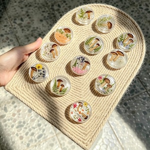 May include: A collection of eleven circular clear resin cabochons, each containing pressed dried mushrooms, small green ferns, and various colorful wildflowers. The pieces are arranged on a textured, light beige woven rope display board.
